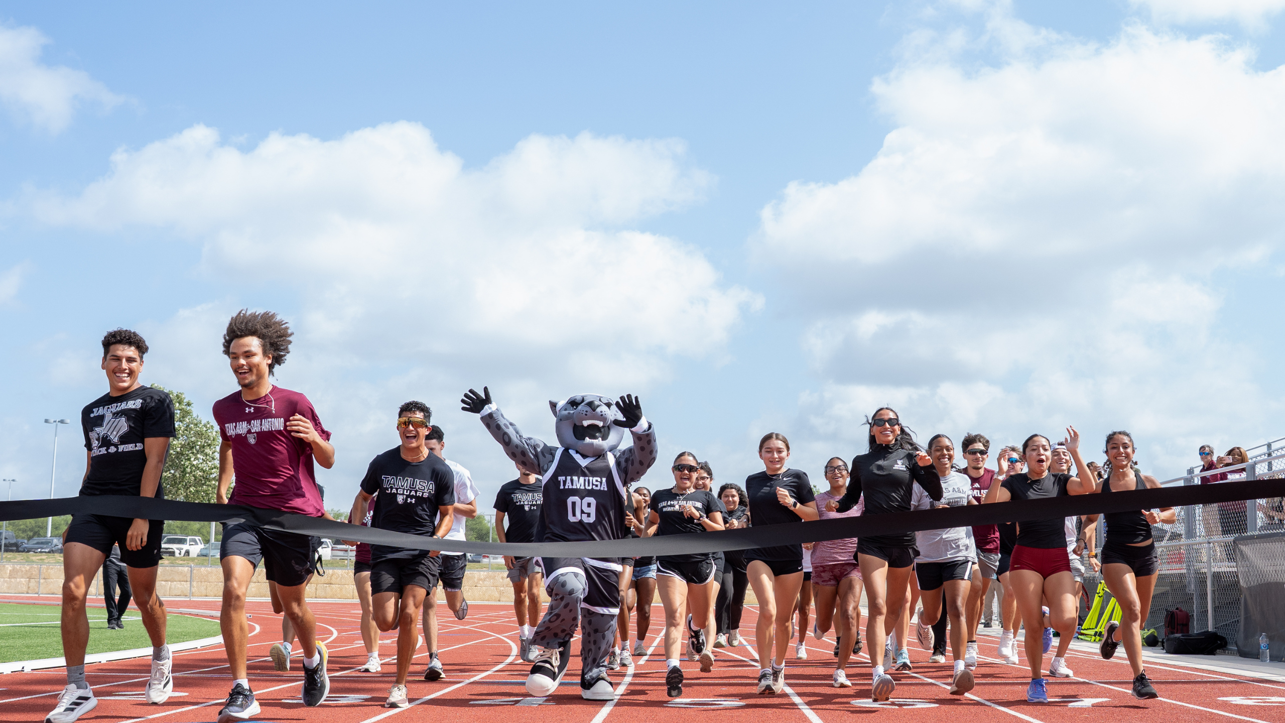 Student-athletes from A&M-San Antonio running through the ribbon at the finish line to simulate a ribbon cutting. 